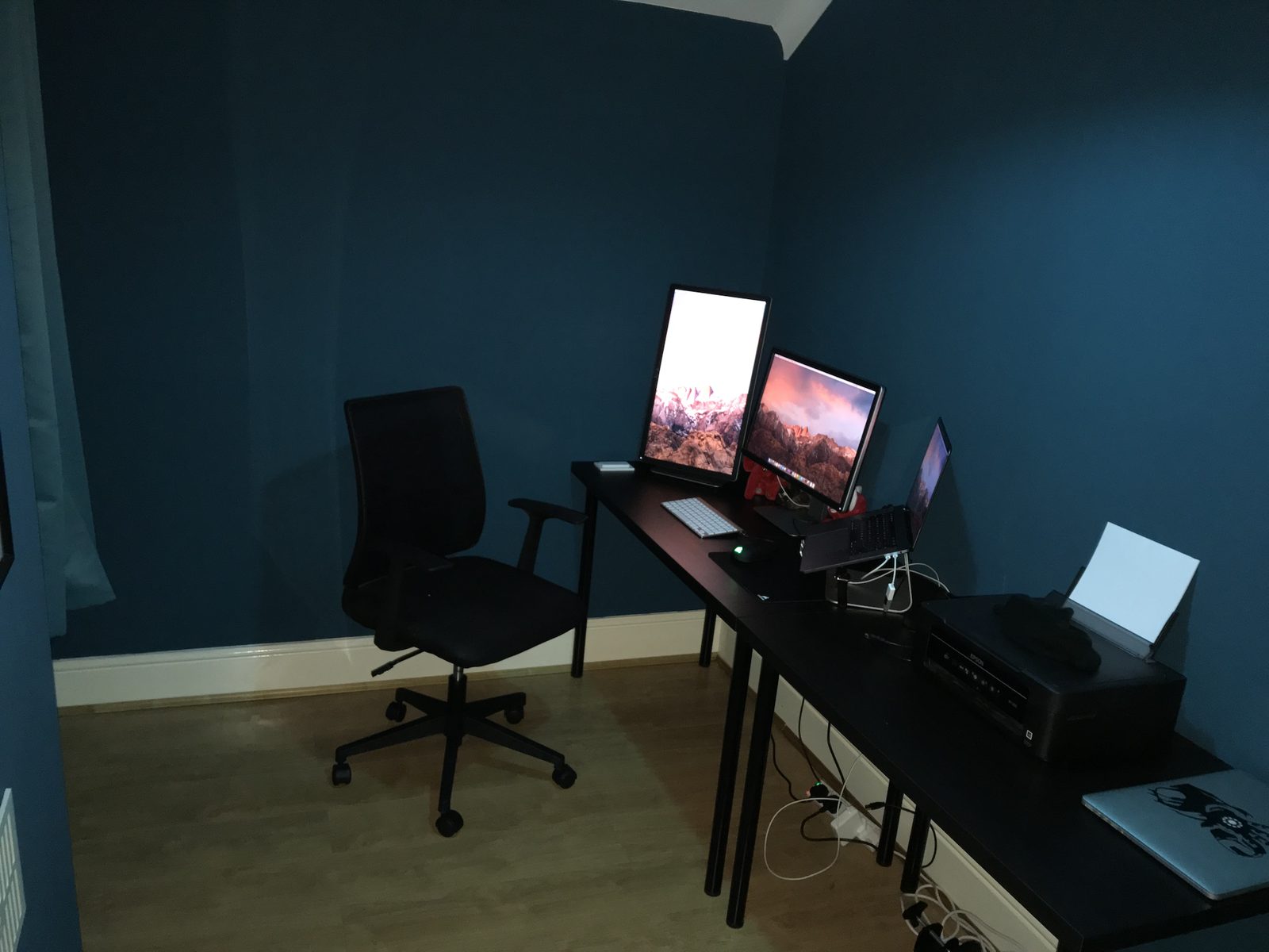 The old office: a dark blue room with two cheap black IKEA desks pushed together, a basic office chair, a portrait and a landscape monitor, a MacBook on a stand and a printer.