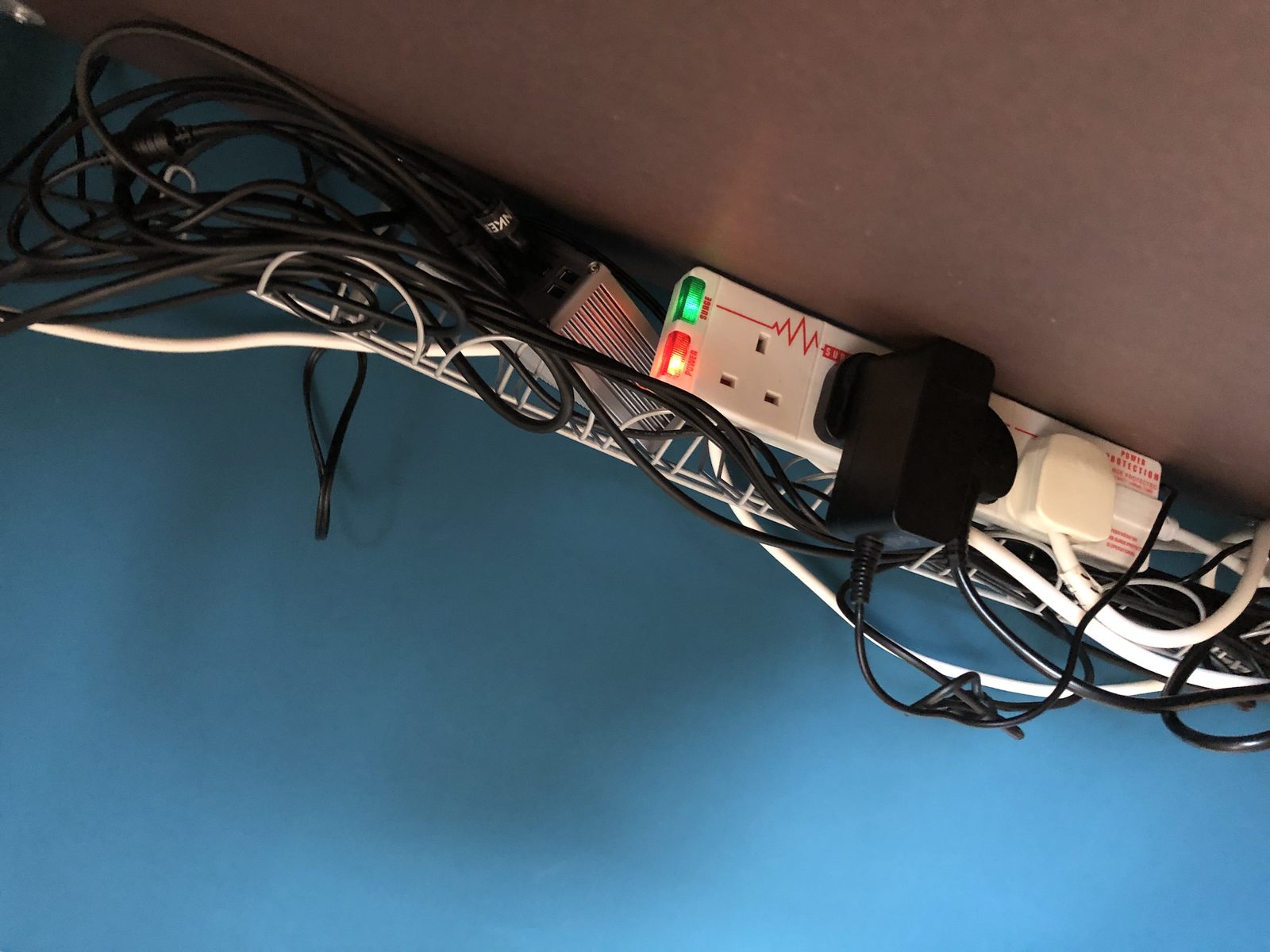 Underneath the desk: a white wire cable tray holding a surge-protected power strip and a tangle of black and white cables.
