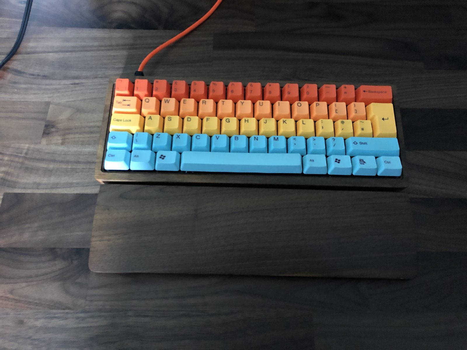 A compact 60% mechanical keyboard in a walnut case with rainbow keycaps in rows of red, orange, yellow and blue, an orange cable and a matching walnut wrist rest.