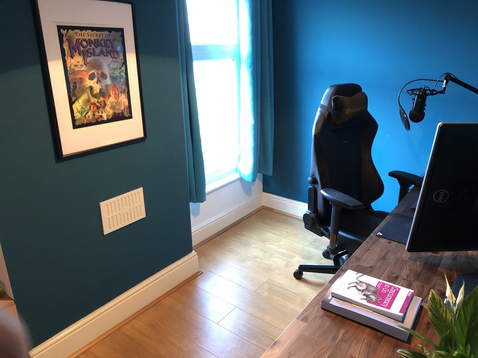 The other side of the room: a framed Secret of Monkey Island poster on a teal wall beside a window with teal curtains, with the chair and microphone at the desk.