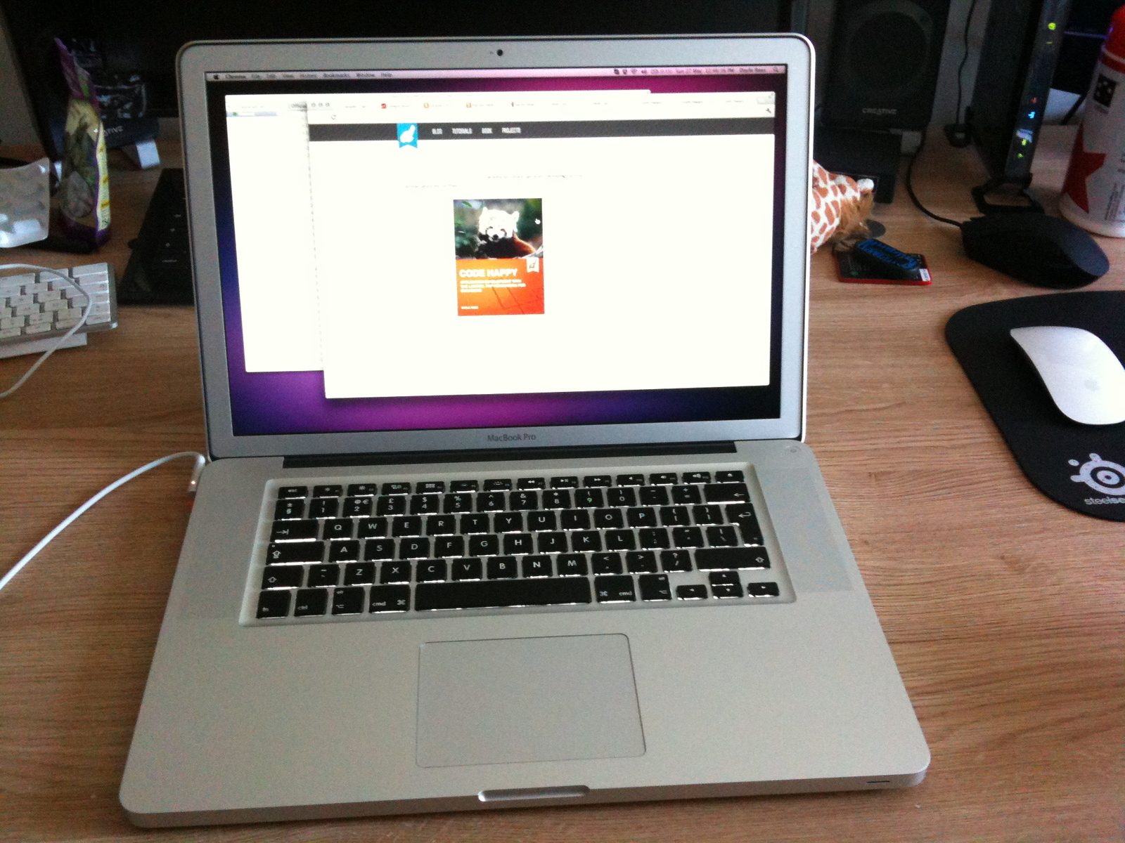 A silver 15-inch MacBook Pro on a wooden desk, its screen showing the Code Happy book cover on an early version of this site.